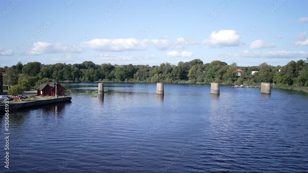 Massive concrete pillars rise from a still Swedish lake, remnants of a missing bridge. A red cabin on a dock that would have led to the bridge. Massive concrete pillars rise from a still Swedish lake, remnants of a missing bridge. A red cabin on a dock that would have led to the bridge.