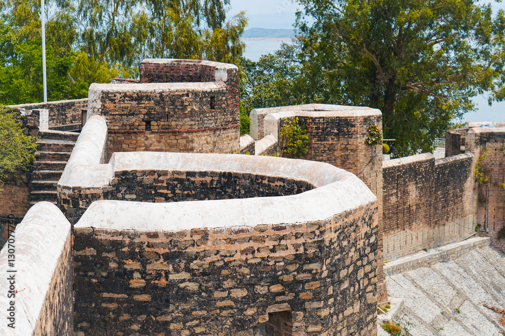 A historic view of Mangla Fort, featuring weathered stone walls and stairways surrounded by lush greenery, with a serene lake in the background. A historic view of Mangla Fort, featuring weathered stone walls and stairways surrounded by lush greenery, with a serene lake in the background.