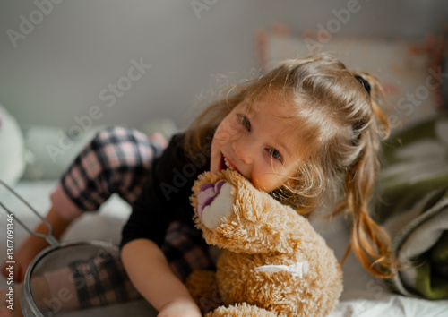 Obraz na plátně Happy little girl playing and chewing her teddy bear in the morning wearing pajamas