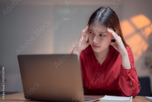 Young woman touching her head and feeling tired while working late at night on her laptop in office, suffering from headache, stress, overwork and sleep deprivation