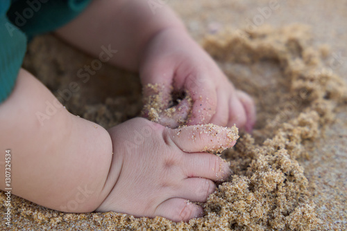 The baby is playing with sand, and the baby's foot is in the sand. The baby has a toothbrush in its mouth, which might be an unrelated object or part of the playtime activity. photo