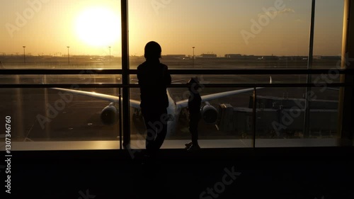 Mother and child stand by terminal window, looking out to plane and airport outdoors during golden sunset. Dramatic lighting and silhouetted appearance. Camera smoothly slide to side, slow motion shot