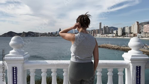 Tourist woman walks up to the white railing to observe expansive Benidorm cityscape and mediterranean coastline at sunny outdoor viewpoint with coastal panorama. Medium shot