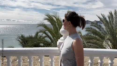 Woman wearing sunglasses and casual clothes looking at the sea from a white balcony with palm trees
