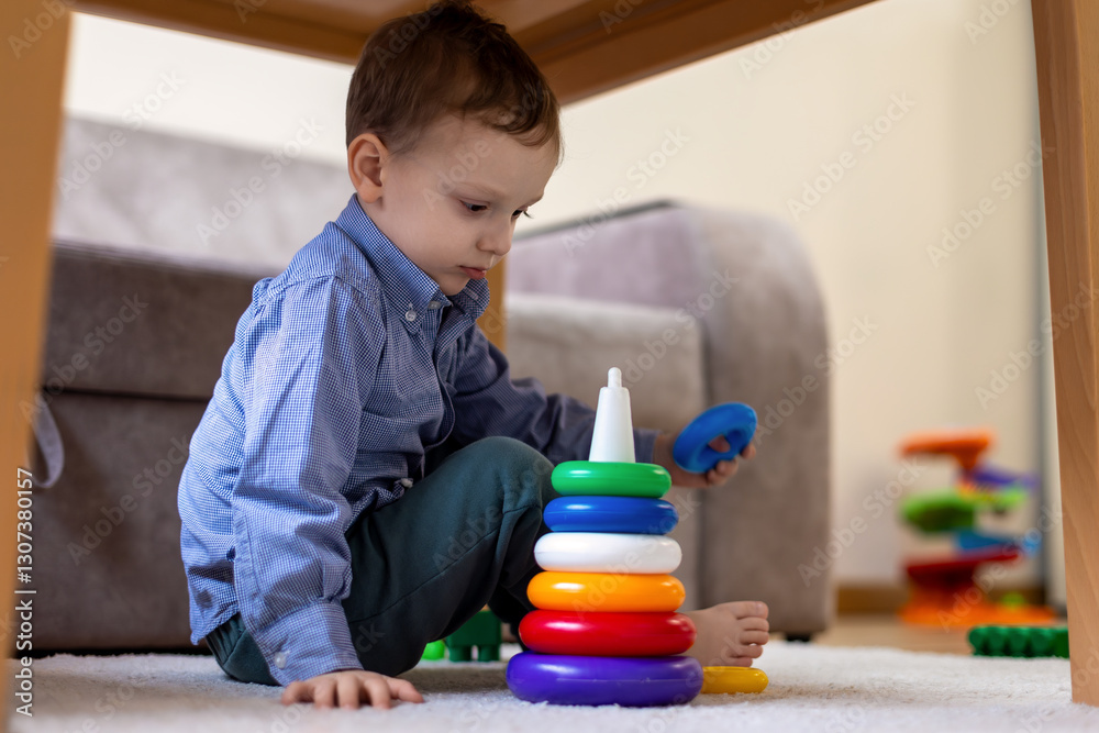 Young boy playing with colorful stacking rings. A young boy sits on the floor under a table, playing with a colorful stacking ring toy. Young boy playing with colorful stacking rings. A young boy sits on the floor under a table, playing with a colorful stacking ring toy.