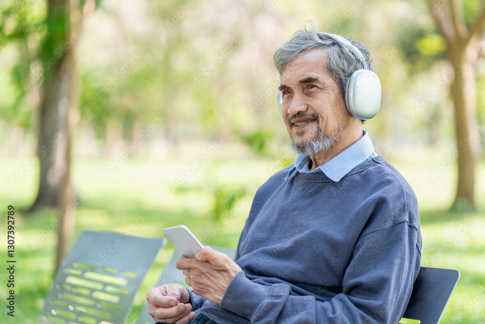 Fototapeta premium portrait happy asian senior man with grey hair and beard wears headset holding smartphone to listening music,sitting on bench in the park