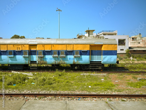 A stationary train carriage sits on a track in an open area. The carriage is painted blue and yellow, with barred windows and metal shutters.