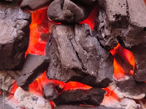 A close-up of burning charcoal with glowing red embers, creating intense heat for grilling and barbecuing. The blackened coals contrast with the fiery glow.