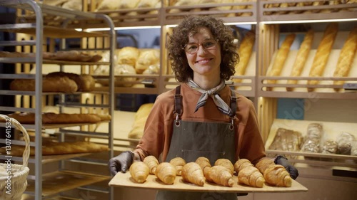 Happy female baker in apron and gloves holding wooden tray filled with freshly baked croissants, smiling at camera in cozy bakery with shelves of bread in background. Video portrait