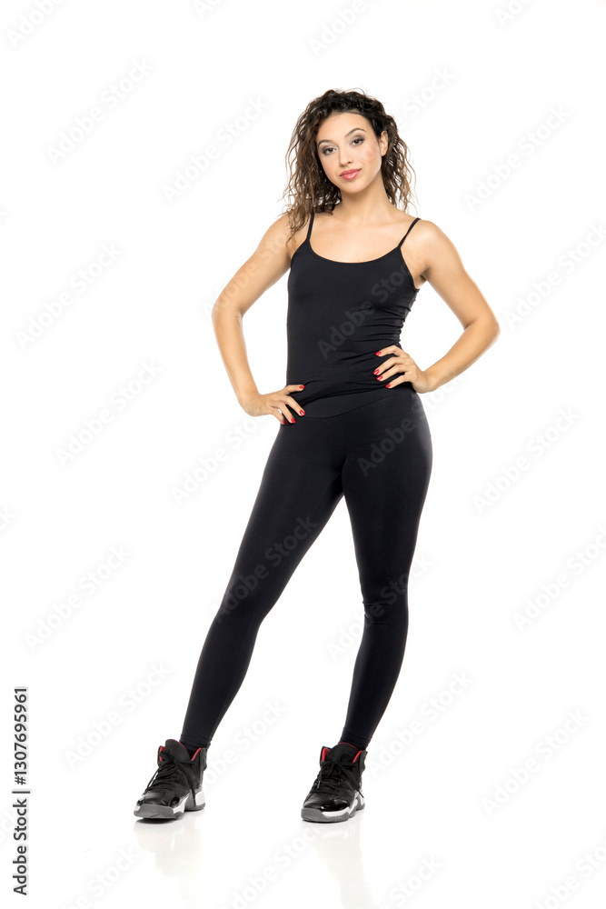 Full-length portrait of young woman with wavy hair wearing black tank top, leggings and athletic shoes, standing confidently against white background Full-length portrait of young woman with wavy hair wearing black tank top, leggings and athletic shoes, standing confidently against white background