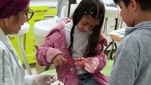 A little girl learning to brush her teeth with a denture model. She finds herself next to another child who is watching her and next to her dentist.