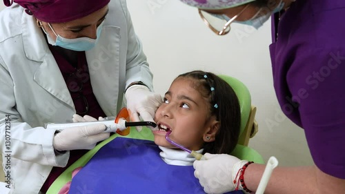 Dentist and nurse examining the teeth of a young girl in a modern dental clinic. The dentist is using a dental curing LED light along with professional medical instruments to check and treat her oral 