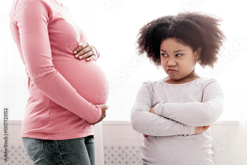 Jealousy In Children Concept. Offended little afro girl looking at pregnant mom's belly with reproach and envy