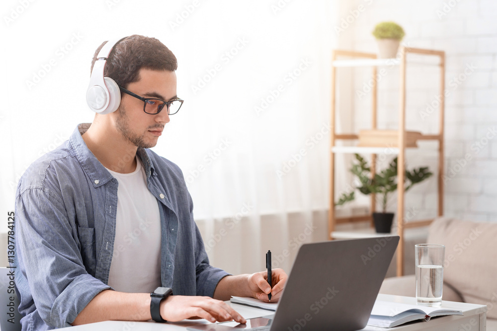 Training and conference online. Busy guy with glasses and headphones makes notes in notebook and works on laptop in living room interior Training and conference online. Busy guy with glasses and headphones makes notes in notebook and works on laptop in living room interior