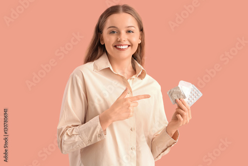 Young woman pointing at contraceptives on pink background