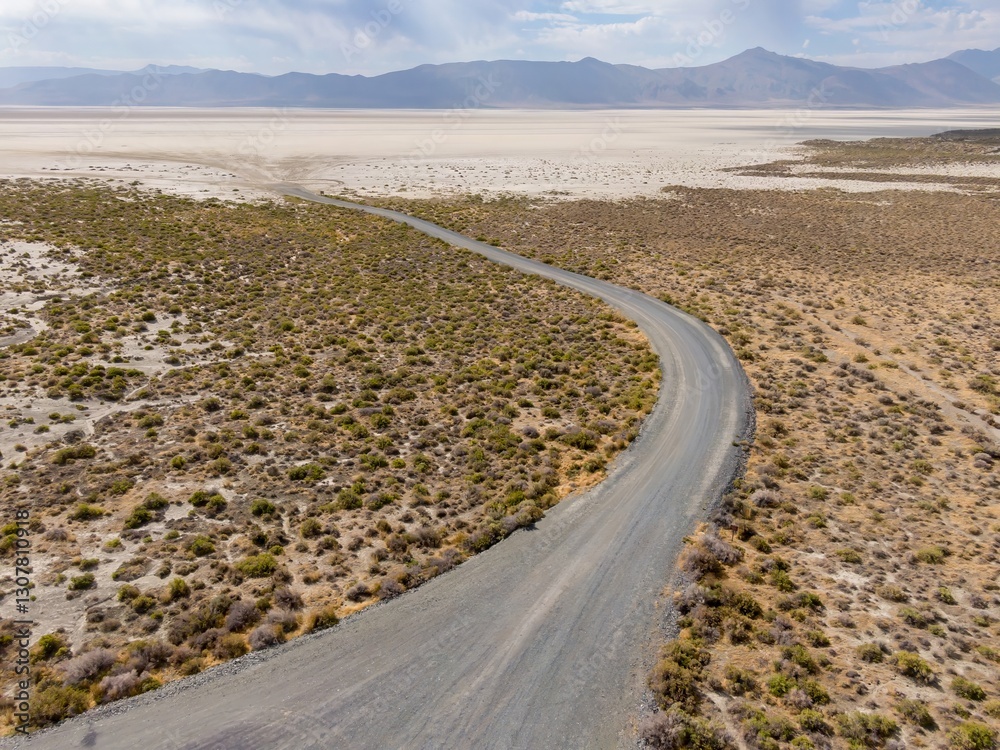 Fototapeta premium A winding gravel road cuts through the arid landscape of the Black Rock Desert in Nevada, USA. The road leads towards a vast, dry lakebed and distant mountains. Gerlach, Nevada, USA