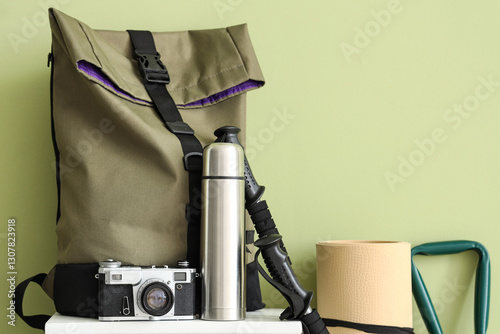 Set of camping equipment with backpack, photo camera and thermos near green wall