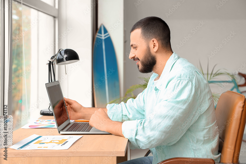 Fototapeta premium Young man sitting at workplace and using laptop for online work at home