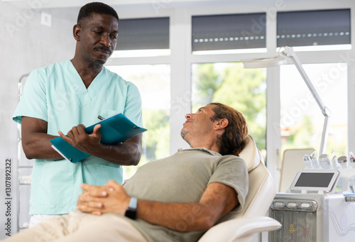 Male patient checks correctness of data entered in clients questionnaire. African man doctor writes missing data in report photo