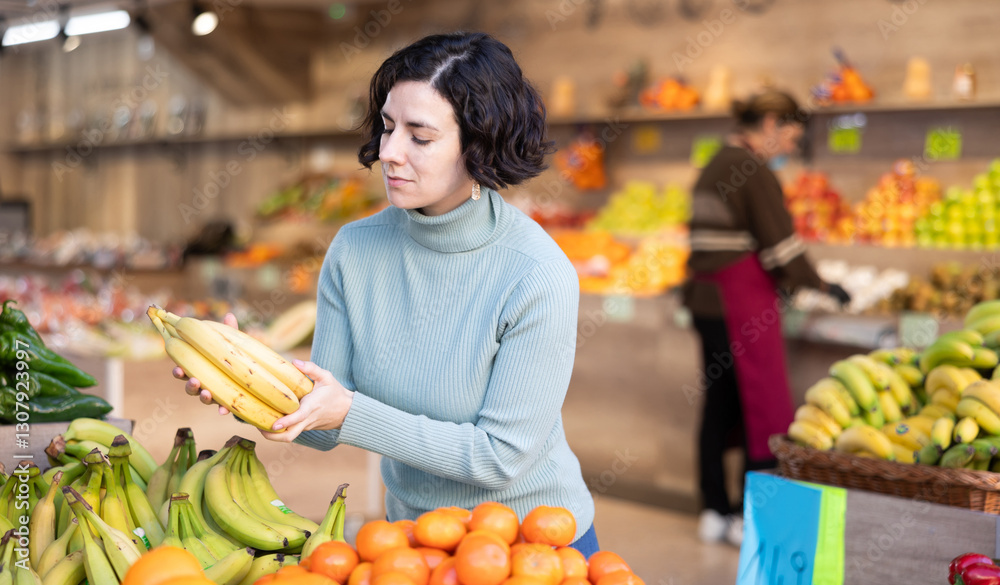 Obraz premium Adult woman buyer choosing bunch of fresh bananas in vegetable shop