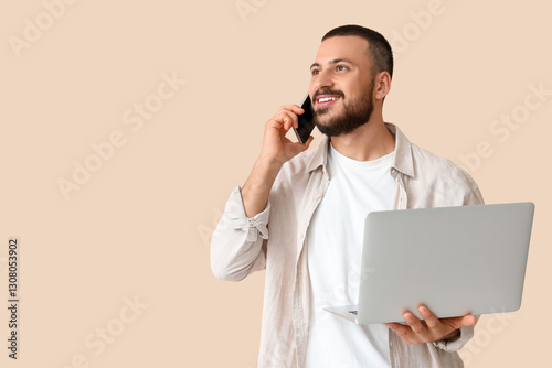 Young man with laptop taking on mobile phone on beige background