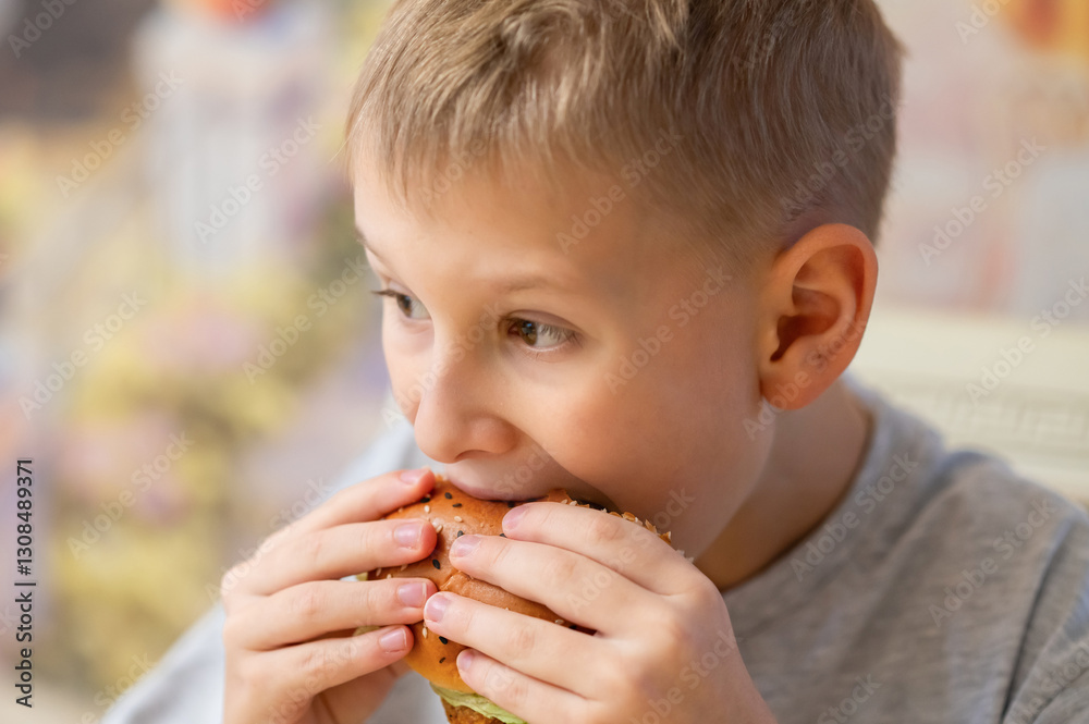 A young boy with short blond hair enjoys a delicious burger, taking a big bite. He holds the sesame seed bun with both hands, savoring the taste. A young boy with short blond hair enjoys a delicious burger, taking a big bite. He holds the sesame seed bun with both hands, savoring the taste.