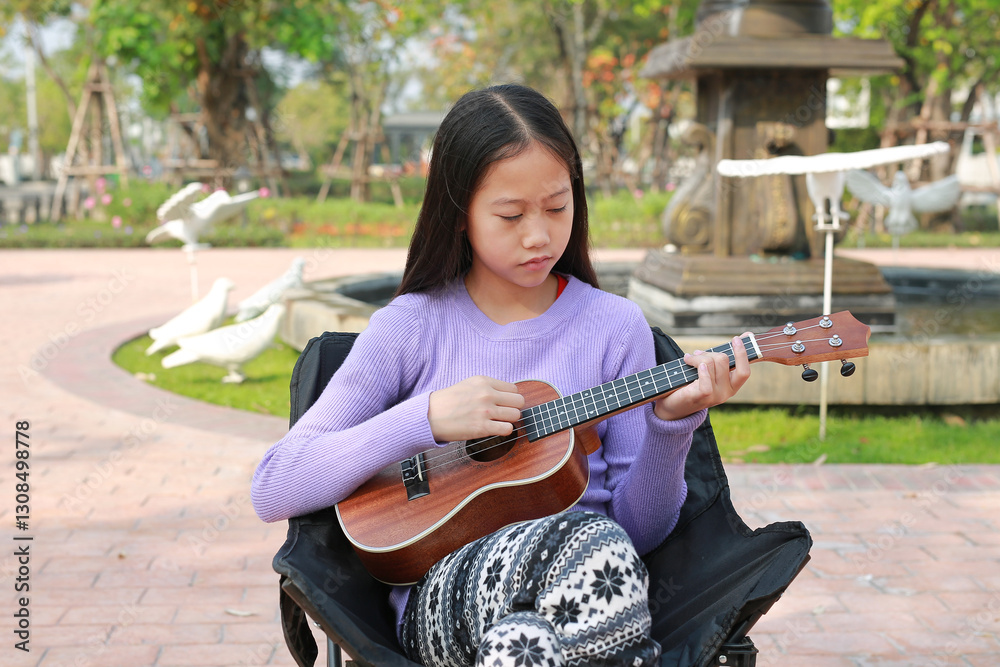 Portrait of Asian pretty girl playing classic ukulele while sitting on canvas chair in the garden.