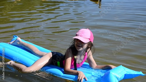 girl in pink cap and swimsuit floating on inflatable blue mattress. Happy kids have fun on the beach swimming circles. summer holidays. Children enjoy vacation and spend outdoors at the seaside