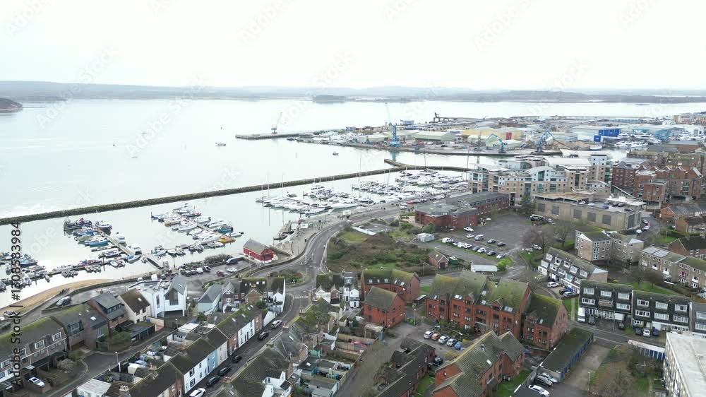 Aerial drone view of coastal town with marina full of boats, residential buildings, and industrial port in distance. Roads curve through town, leading to waterfront under overcast sky.