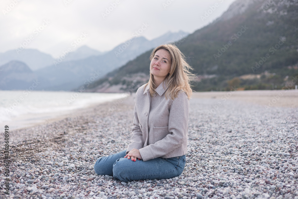 Blonde woman sits on the pebbled beach in winter Blonde woman sits on the pebbled beach in winter