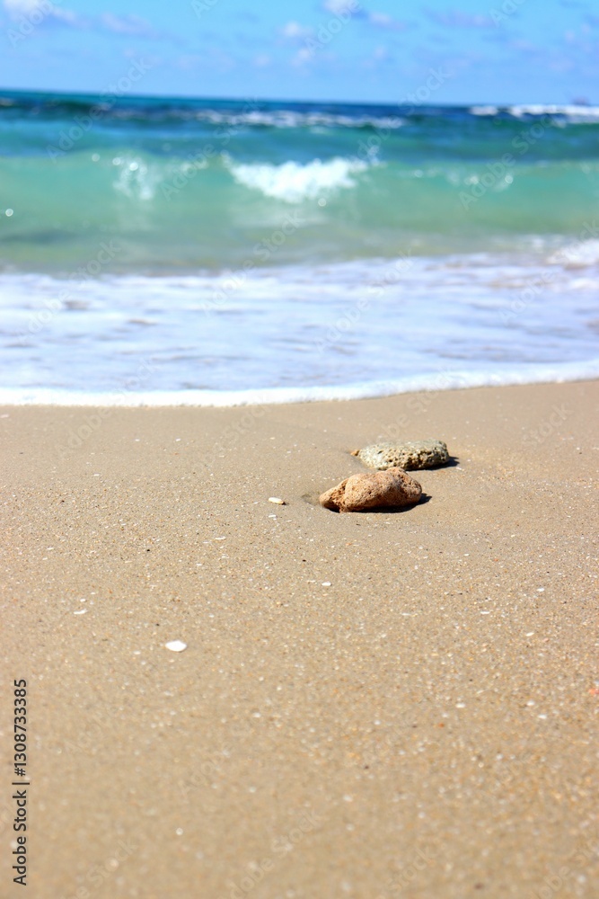 Two rocks on a sandy beach, with turquoise waves and a blue sky in the background. Two rocks on a sandy beach, with turquoise waves and a blue sky in the background.