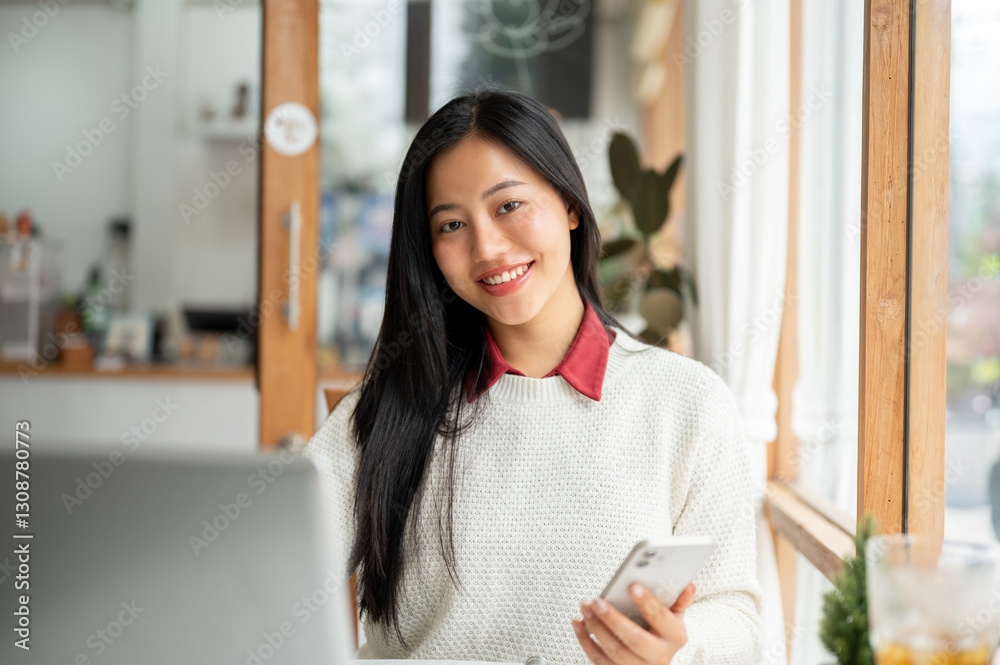 A smiling asian woman is holding the smartphone while looking straight into camera, Sitting with a laptop in the cafe. A smiling asian woman is holding the smartphone while looking straight into camera, Sitting with a laptop in the cafe.