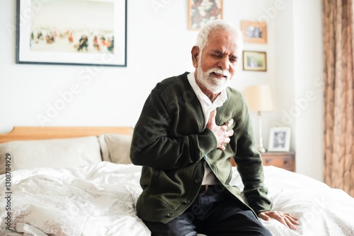 Elderly Indian man sitting on bed, clutching chest in discomfort. Senior male experiencing chest pain. Concerned elderly man in bedroom setting. Elderly Indian man unwell with chest pains.