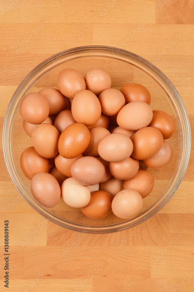 A top-down view of a clear glass bowl filled with brown eggs, set against a light wooden surface A top-down view of a clear glass bowl filled with brown eggs, set against a light wooden surface