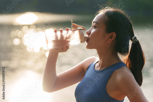 Young sportswoman drinking water from a bottle while standing by a serene lake at sunset, savoring a refreshing break after an invigorating workout in the golden hour light