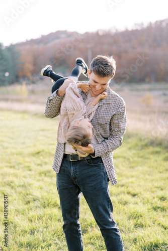 Smiling dad holding laughing little girl upside down on shoulder in meadow