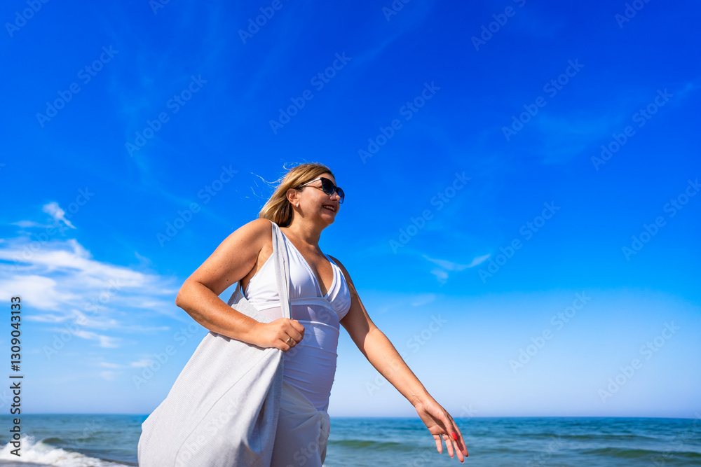 Summer vacation by sea. Beautiful elegant middle aged woman tourist with beach bag walking along sea on sunny day. Side view Summer vacation by sea. Beautiful elegant middle aged woman tourist with beach bag walking along sea on sunny day. Side view