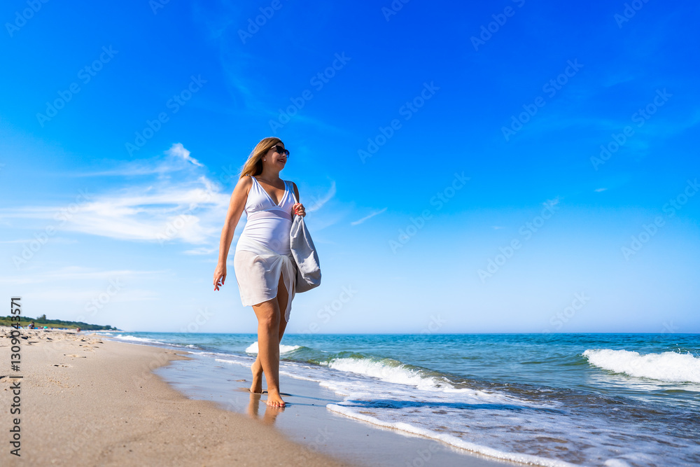 Summer vacation by sea. Beautiful elegant middle aged woman tourist with beach bag walking along sea on sandy beach on sunny day. Front view Summer vacation by sea. Beautiful elegant middle aged woman tourist with beach bag walking along sea on sandy beach on sunny day. Front view