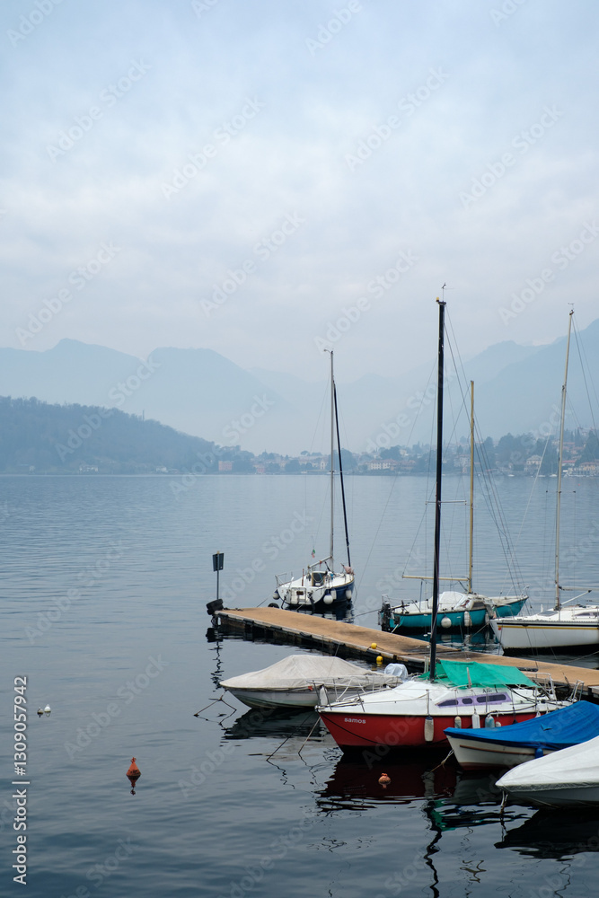 Fototapeta premium Tranquil lakeside marina with moored sailboats, calm water reflections and misty mountain backdrop creating a peaceful nautical atmosphere