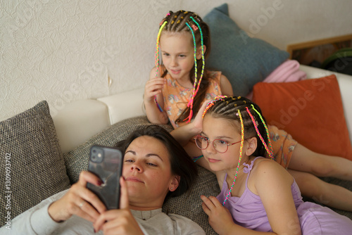 A mother lying on the couch, scrolling through her smartphone, with smiling children beside her. A concept of modern digital life and family.