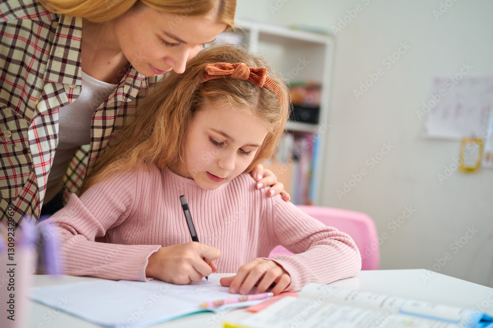 Mother helping daughter with homework at desk, looking to workbook and explaining tasks in a supportive and engaging way Mother helping daughter with homework at desk, looking to workbook and explaining tasks in a supportive and engaging way