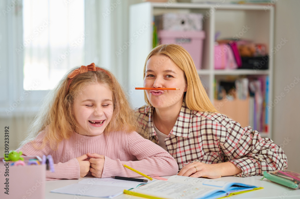 Playful mother balancing pencil on her lip makes funny faces as her laughing daughter works, creating joyful prep scene filled with love and silliness Playful mother balancing pencil on her lip makes funny faces as her laughing daughter works, creating joyful prep scene filled with love and silliness