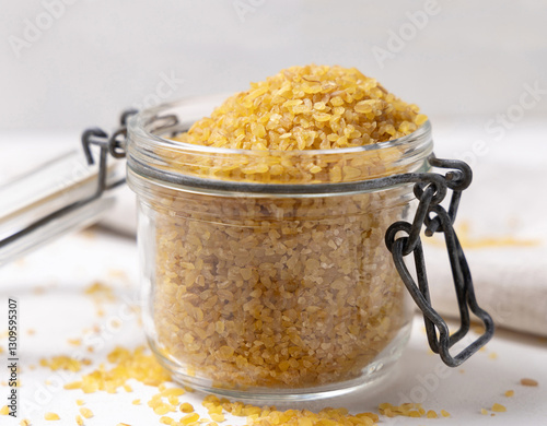 Opened glass jar of raw dry uncooked yellow bulgur grain on white table cloesup. Ancient food photo