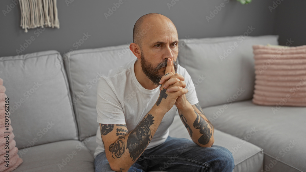 Hispanic man with a bald head and beard, sitting thoughtfully in a modern living room, wearing a white t-shirt, and showcasing tattoos on both arms. Hispanic man with a bald head and beard, sitting thoughtfully in a modern living room, wearing a white t-shirt, and showcasing tattoos on both arms.