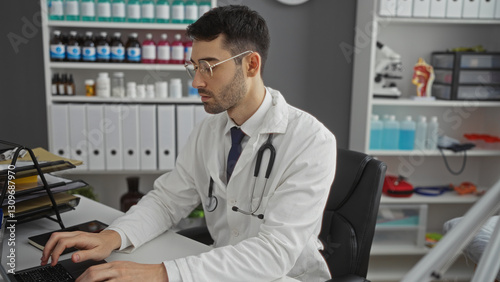 Fotografía Young man working in a hospital clinic room, wearing a lab coat with stethoscope