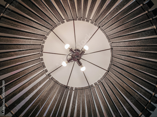 A close-up view of a modern chandelier with multiple exposed light bulbs, surrounded by a circular geometric structure. The design features radial wooden or metal slats.