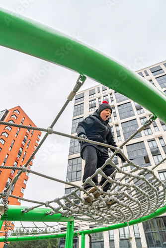 A child in warm clothes climbing a rope net on a playground surrounded by modern apartment buildings. Concept of outdoor activity in urban spaces