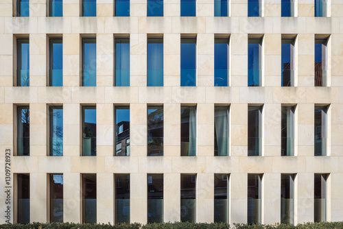 A contemporary building exterior featuring a beige stone facade with a repetitive pattern of narrow, rectangular windows.