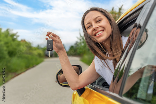 Obraz na płótnie A cheerful woman in a white sleeveless shirt holds car keys while leaning out of the window of a yellow car on a sunny day, surrounded by a scenic countryside road. w salonie