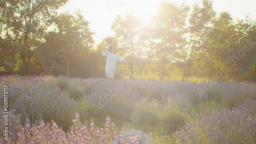 Woman in a white lace dress and hat running through a lavender field at sunset, surrounded by lush greenery and warm golden sunlight. Romantic summer scene. Nature and relaxation concept.